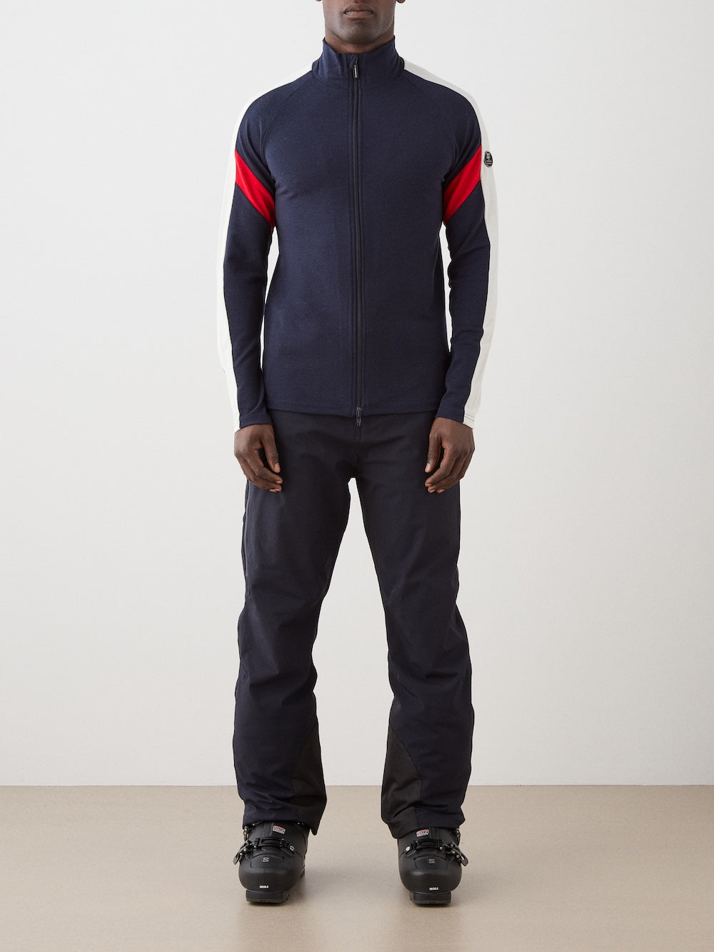 A person is wearing the We Norwegians Zermatt FullZip men jacket with red and white sleeve stripes, paired with dark pants and black ski boots, standing against a plain background.