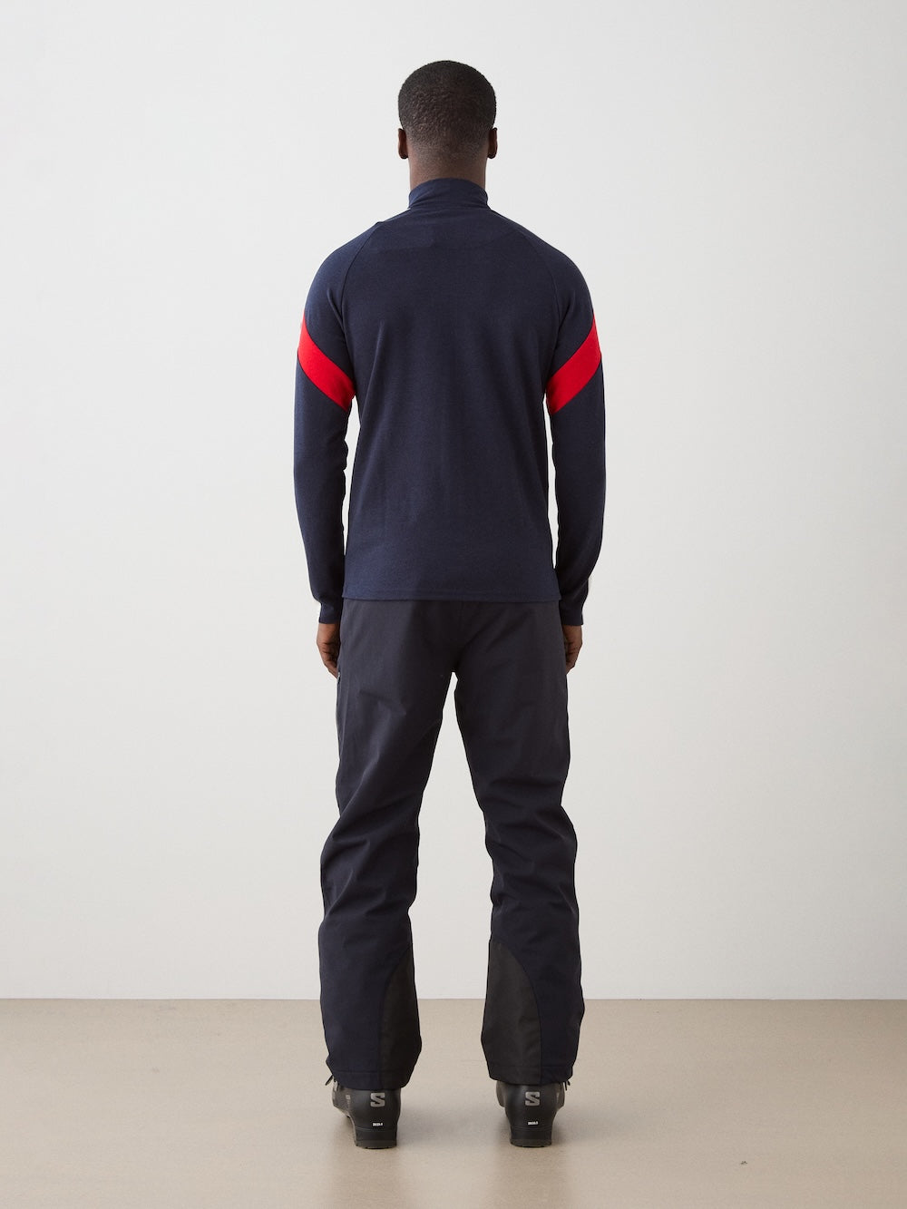 A person stands facing away, wearing the We Norwegians Zermatt FullZip men in navy with a red arm stripe and dark pants, in a minimal indoor setting with a light floor and plain white wall.