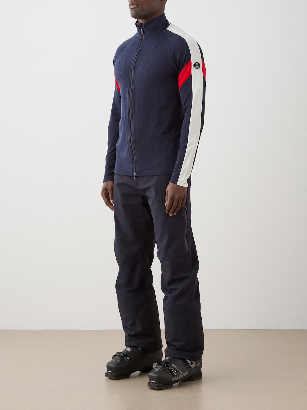 A man indoors wears the We Norwegians Zermatt FullZip men jacket in navy with white and red sleeve stripes, paired with navy pants and black ski boots. The ski mid-layer stands out against the plain, light background.
