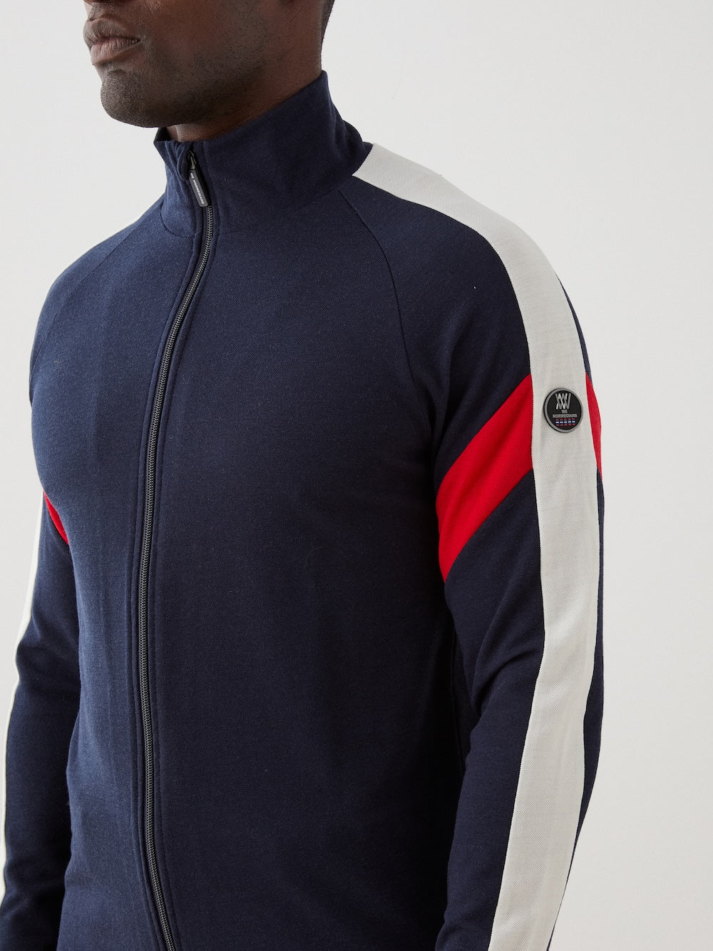 A man wears the We Norwegians Zermatt FullZip men’s jacket in navy blue, featuring white and red sleeve stripes and a small circular logo patch on the upper arm. Only his upper body and lower face are visible, showcasing this sporty ski mid-layer.