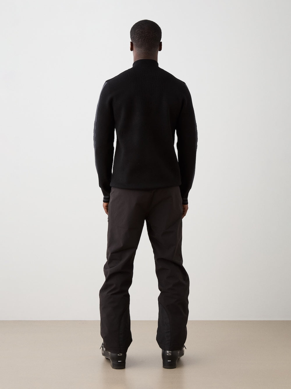 A person faces away from the camera, wearing We Norwegians' Rocky ZipUp men sweater and matching pants against a white wall and beige floor, evoking vintage alpine style.