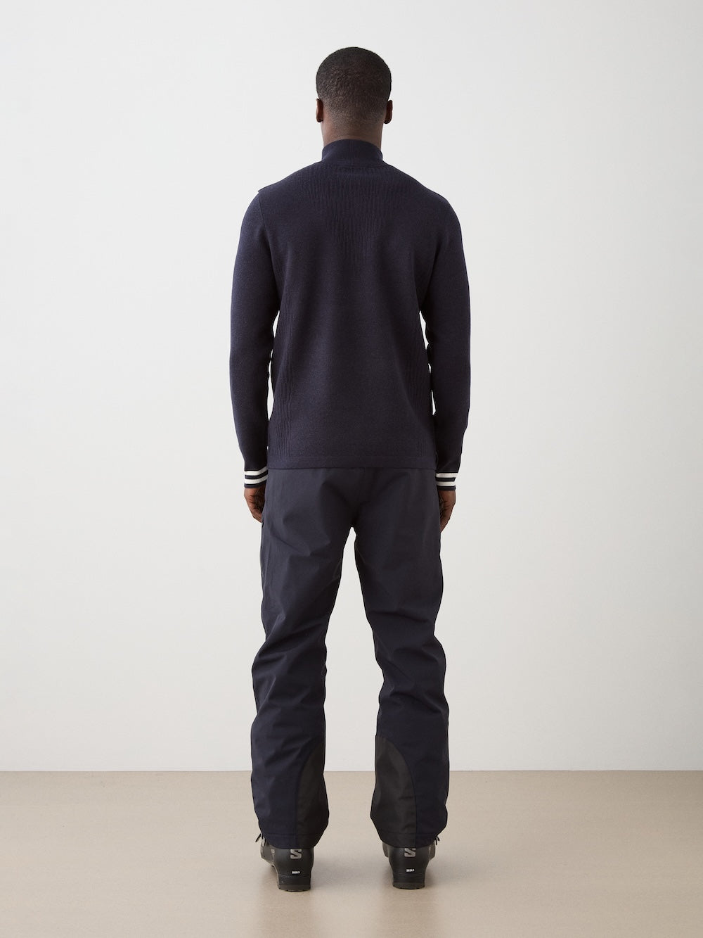A person wears the We Norwegians Rocky ZipUp men’s dark long-sleeve top with black shoes, channeling vintage alpine style against a plain light wall and beige floor, standing facing away from the camera.