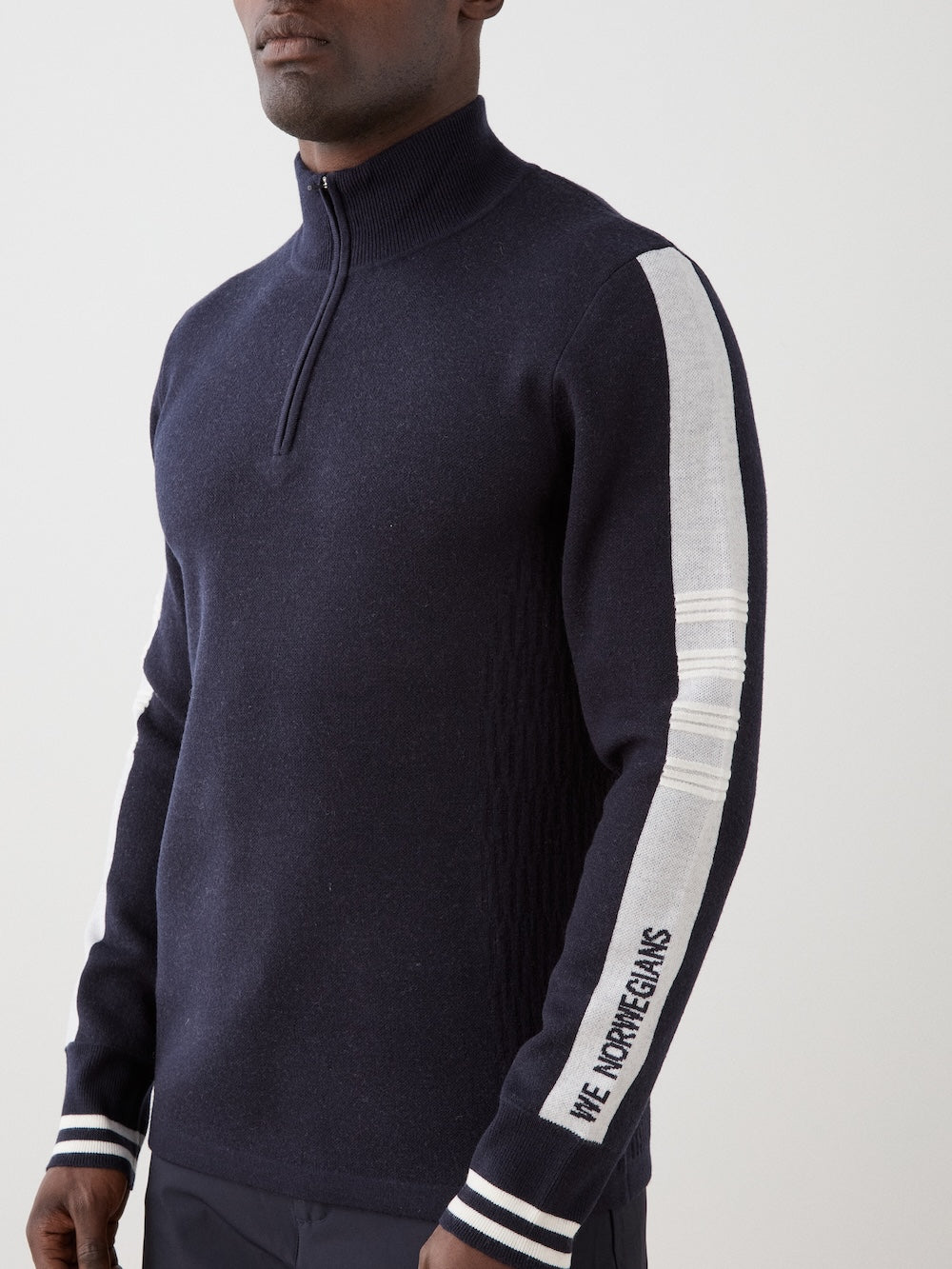 A man wearing the We Norwegians Rocky ZipUp men’s sweater—navy blue with white and gray sleeve stripes and "WE NORWEGIANS" on the left sleeve—stands against a plain light background, highlighting vintage alpine style.