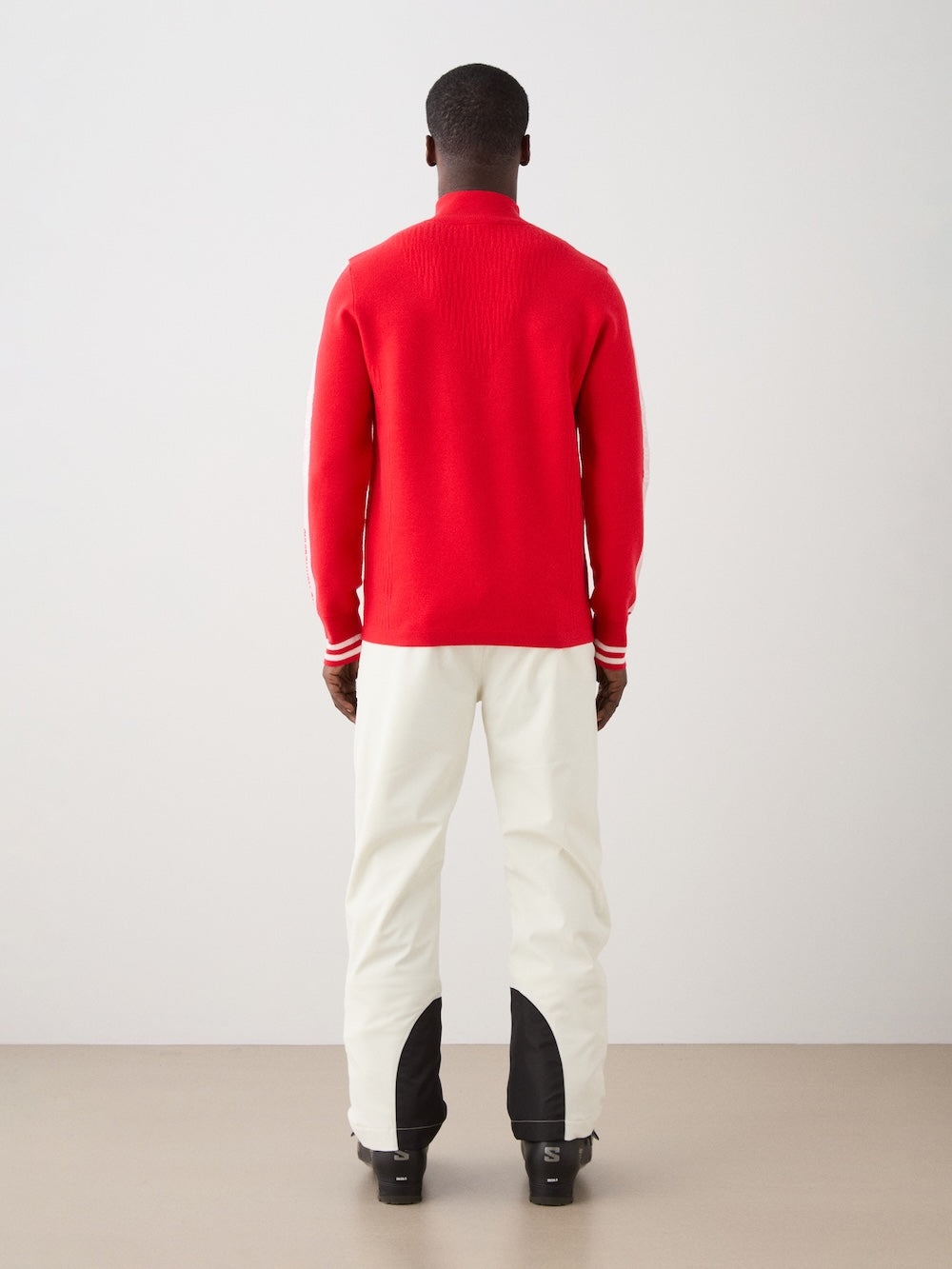 A person stands with their back to the camera, wearing the bright red We Norwegians Rocky ZipUp men sweater and off-white pants with black leg accents, set against a plain indoor backdrop.