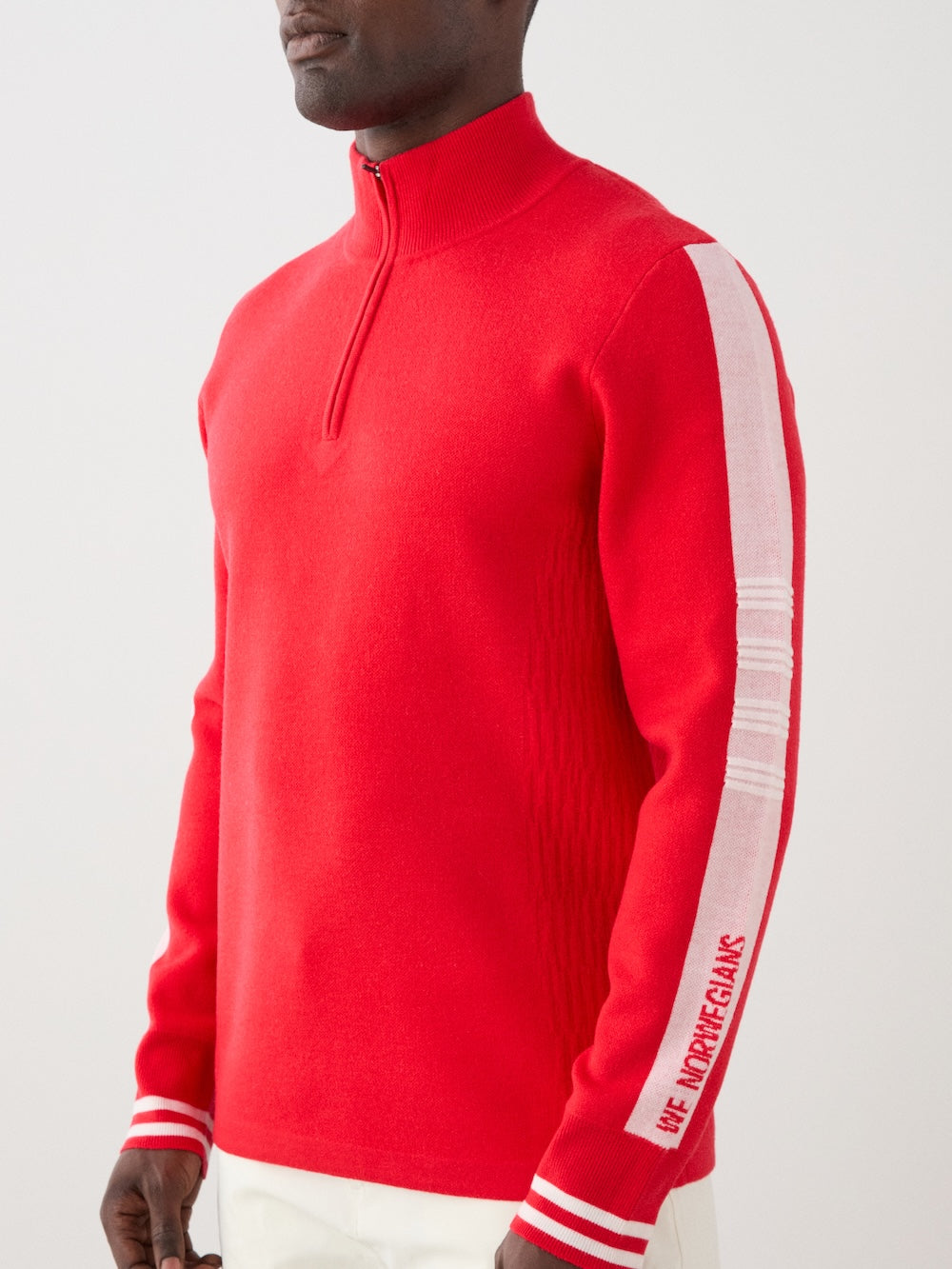 A person wearing the We Norwegians Rocky ZipUp men sweater, featuring a bright red color with white sleeve stripes and text, styled with white pants, evokes vintage alpine vibes against a plain light backdrop.