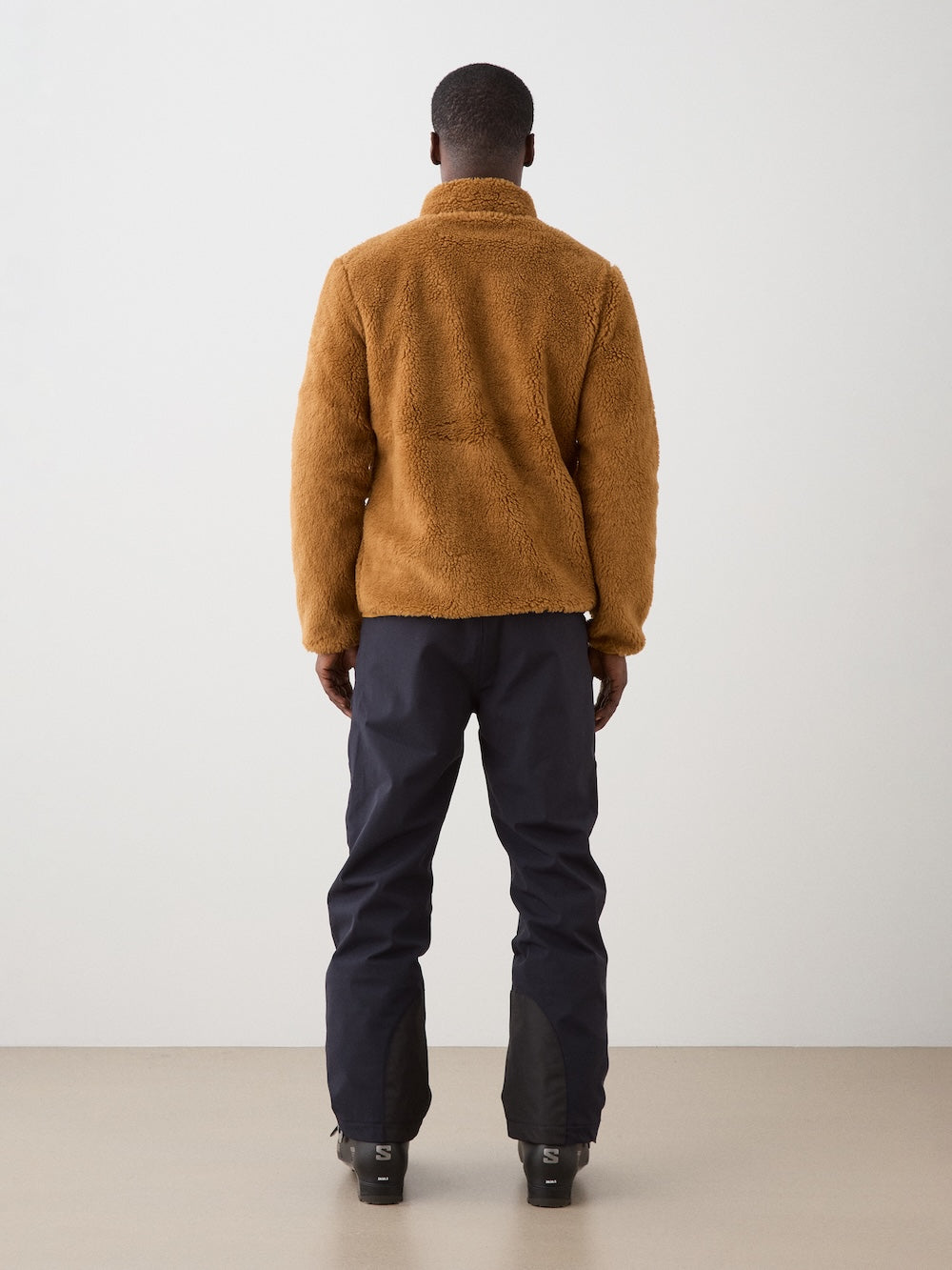 A man faces away from the camera wearing the We Norwegians Kebnekaise Jacket for men, styled with dark pants and black boots against a simple light background.
