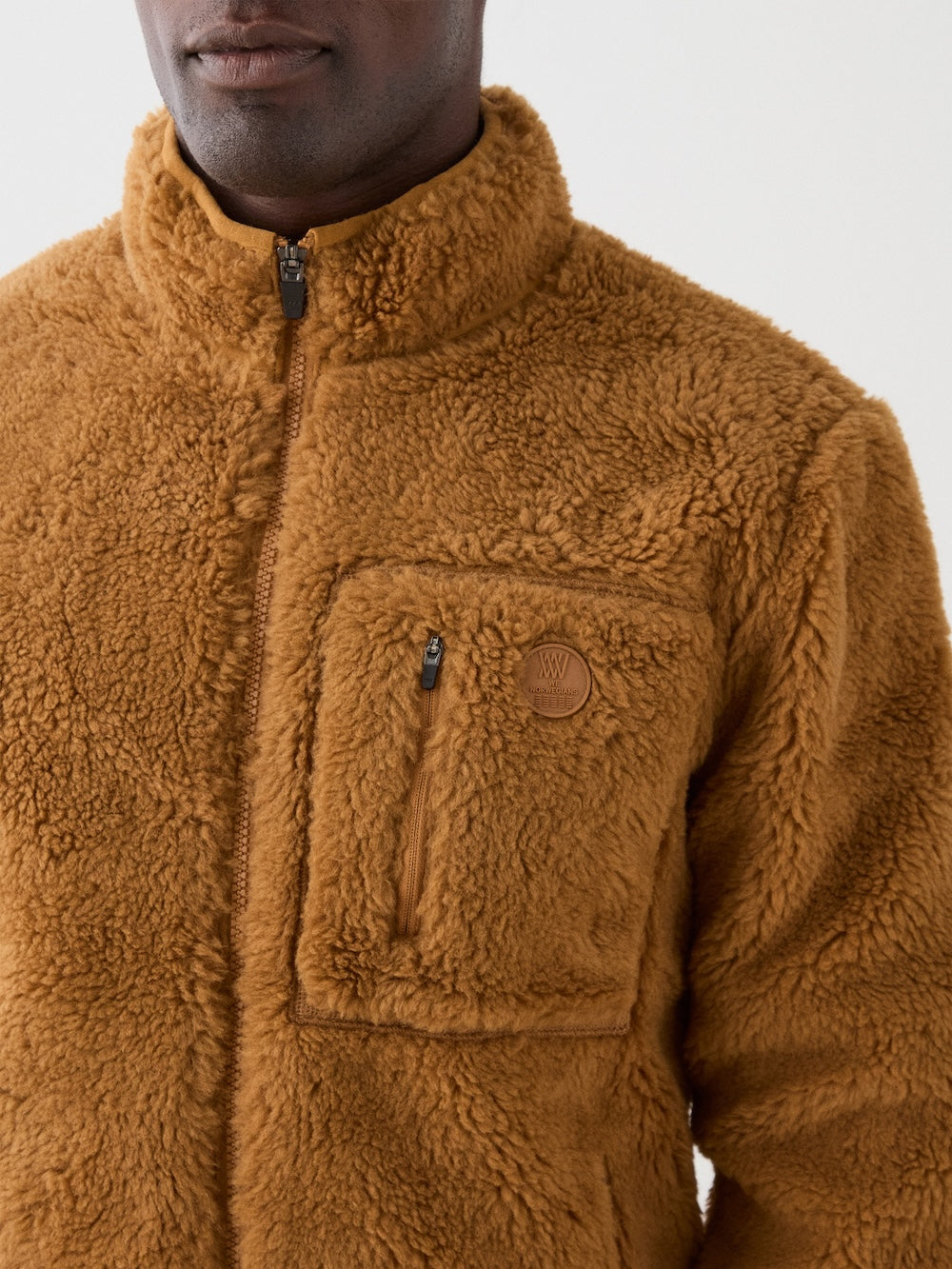 A man wears the We Norwegians Kebnekaise Jacket—a brown, fuzzy, wool-blend zip-up with high collar and zippered chest pocket—shown from the upper torso against a plain background, ideal as a cozy ski mid-layer.