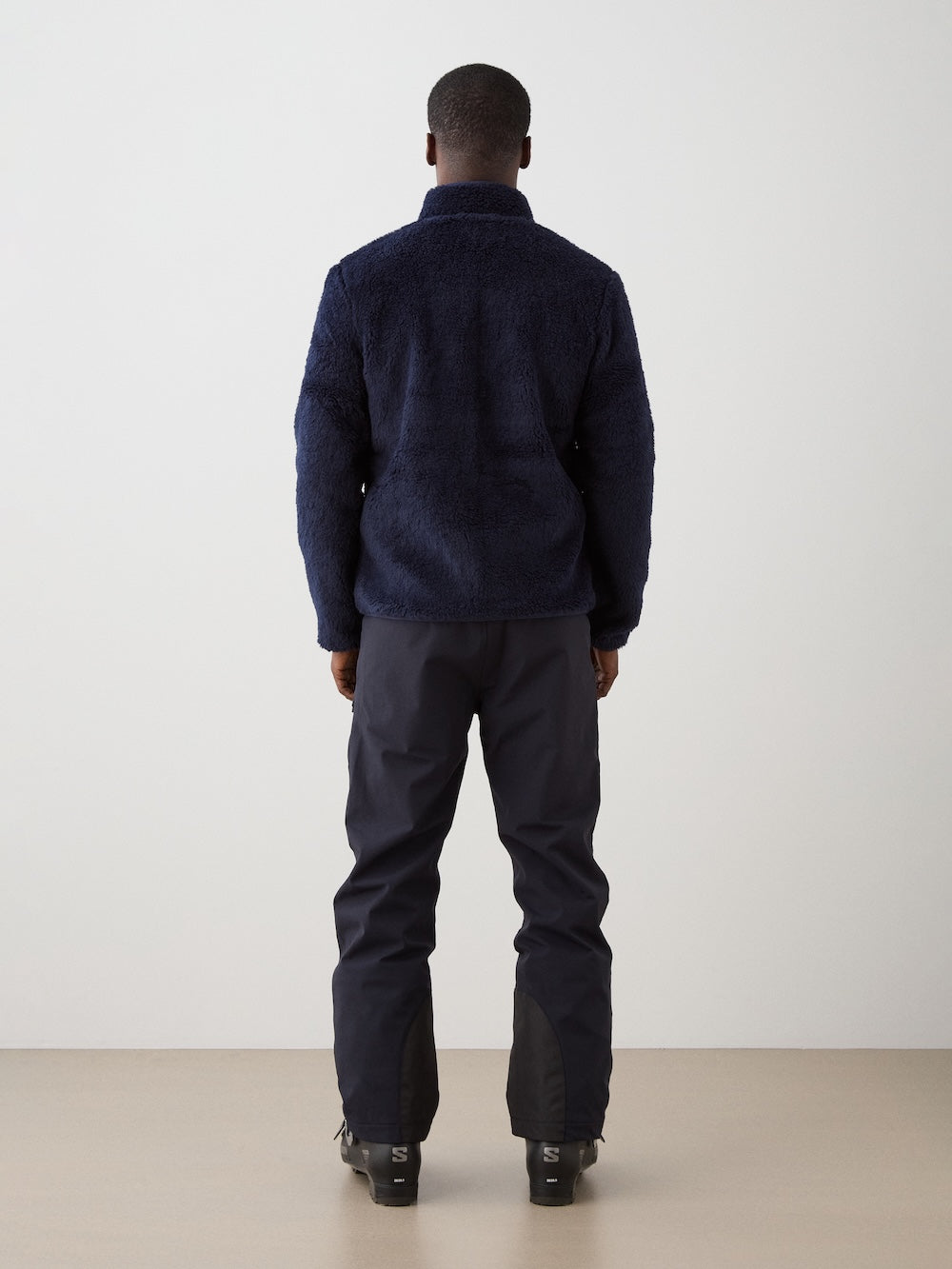 A person faces away from the camera wearing We Norwegians' Kebnekaise Jacket men in dark navy, paired with black pants and ski boots, standing against a plain white wall and beige floor.
