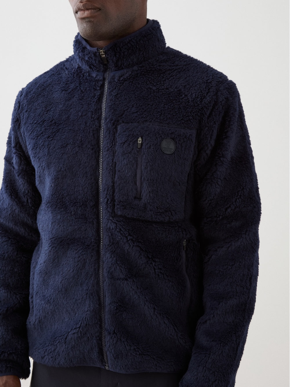 A man wearing the We Norwegians Kebnekaise Jacket—a navy zip-up fleece mid-layer with a chest pocket and stand-up collar—stands against a plain light background.