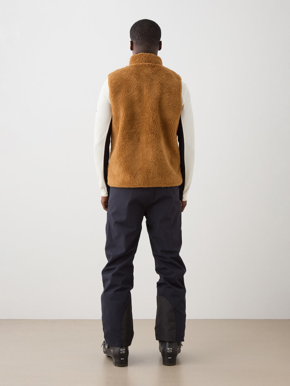 A person stands facing away, wearing the We Norwegians Kebnekaise Vest men—a brown wool-blend pile vest—over a white long-sleeve shirt, dark pants, and black boots, set against a plain light background.