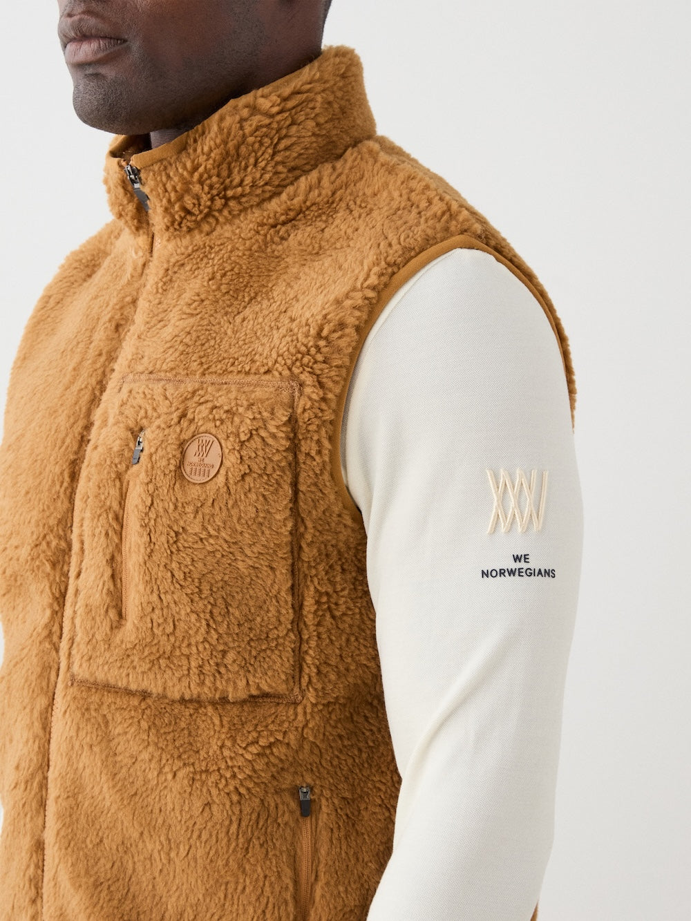 A man wears the We Norwegians Kebnekaise Vest men in tan wool-blend pile over a cream long-sleeve shirt with "WE NORWEGIANS" and "XXVI" on the sleeve—captured from the torso up in profile for a classic après ski mid-layer look.