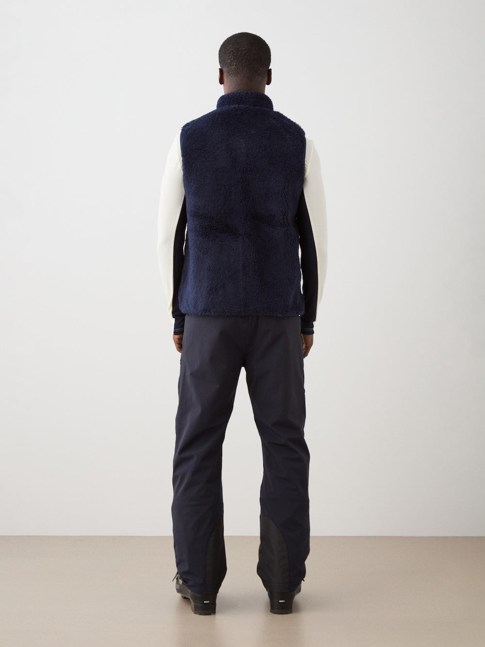 A person faces away from the camera wearing a We Norwegians Kebnekaise Vest men in navy blue over a white long-sleeve shirt and dark pants, standing indoors against a plain background.