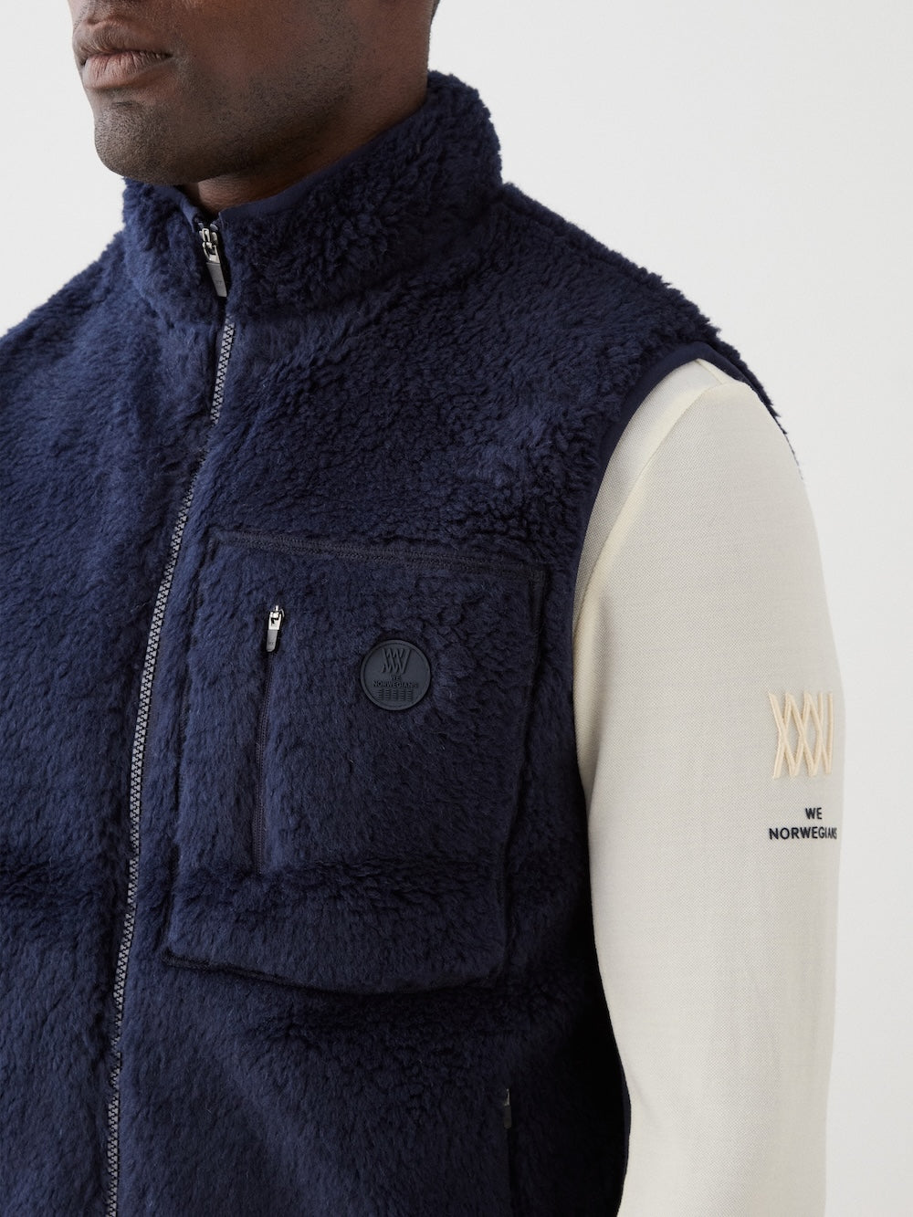 A person wears the We Norwegians Kebnekaise Vest men in navy blue—an ideal wool-blend pile après-ski mid-layer—over a cream long-sleeve shirt with beige Roman numerals on the sleeve. The vest features a chest pocket with a round black patch.