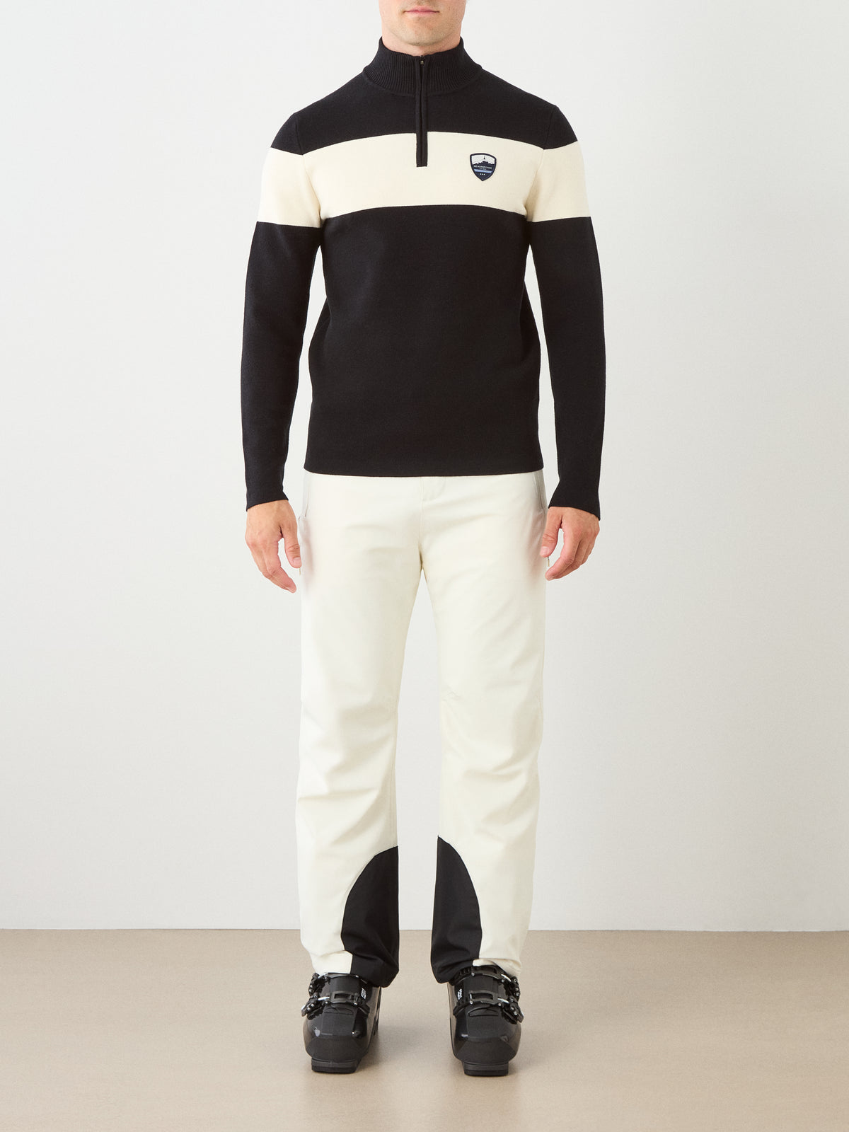 A person standing indoors, facing forward against a plain background, is wearing the Holmenkollen ZipUp men ski sweater by We Norwegians as part of a black and white ski outfit with black ski boots.