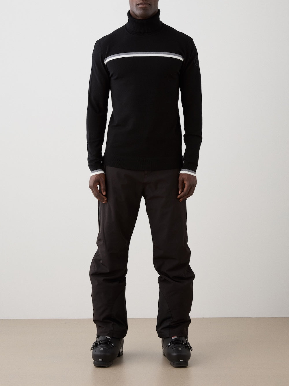 Person wearing a black turtleneck with white and grey stripes on the chest and pants with on a light background