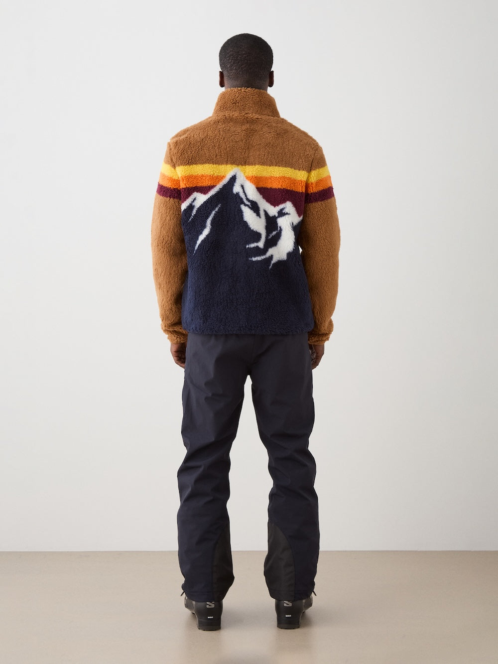 A person stands facing away, wearing the We Norwegians Snowmass Jacket men featuring a wool-blend pile with a mountain and sunset stripe design, paired with dark pants and black boots against a plain light background.