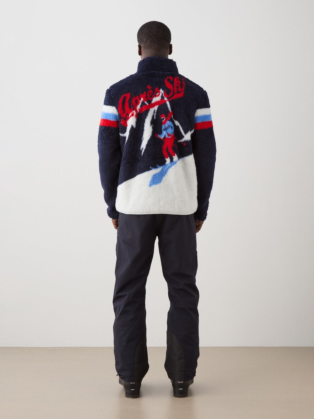 A person faces away in dark pants and the We Norwegians Snowmass Jacket men—an après ski wool-blend jacket with a retro skier and "Après Ski" text on the back. The background is plain and neutral.