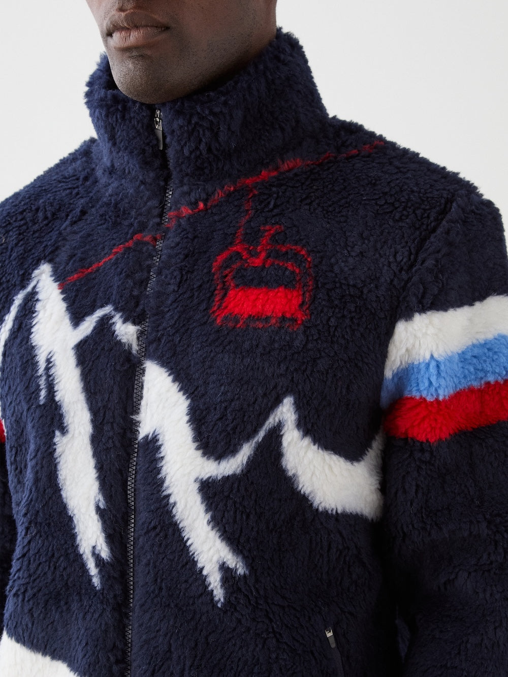 A person in the We Norwegians Snowmass Jacket men, a navy wool-blend fleece with white zigzags, red and blue sleeve stripes, and a red cable car motif on the chest, zipped up to the top.