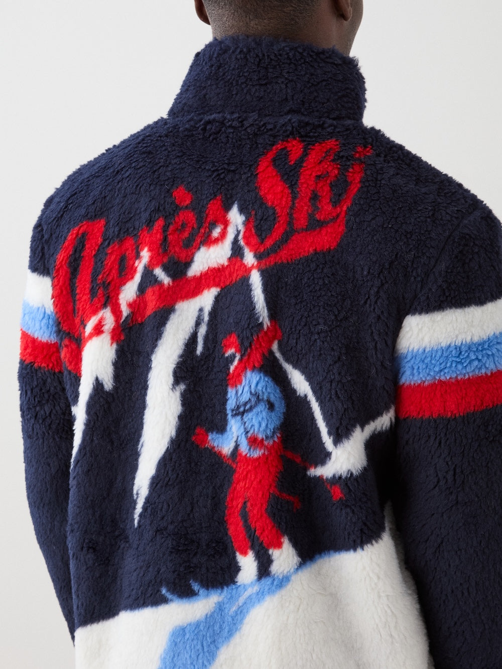 A man wears the We Norwegians Snowmass Jacket men in navy blue wool-blend pile, detailed with red and white stripes and "Après Ski" plus a skier and mountain peaks on the back—an iconic après ski piece.