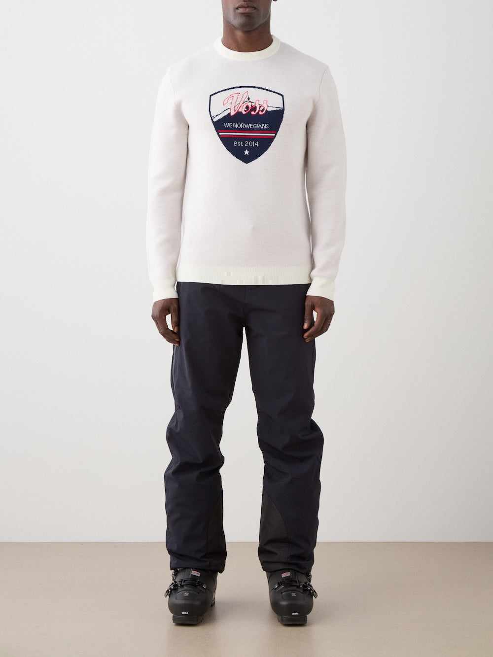 A person stands against a plain background wearing a We Norwegians AfterSki CrewNeck men sweater with a ski graphic, dark pants, and black ski boots.