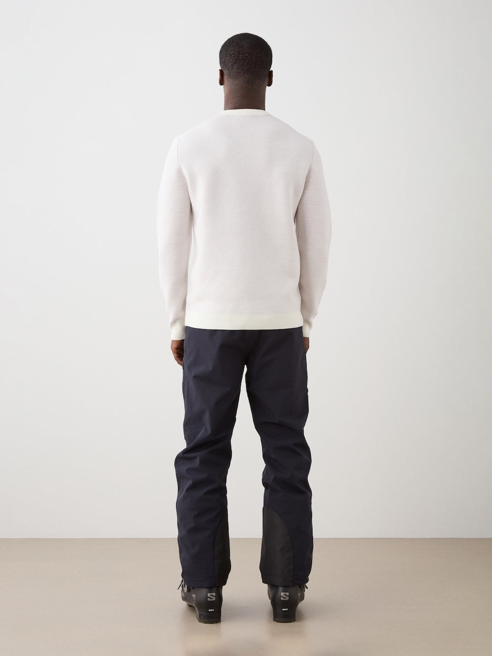 A person faces away from the camera wearing the We Norwegians AfterSki CrewNeck men sweater in white, paired with black pants and black shoes, set against a plain light background.