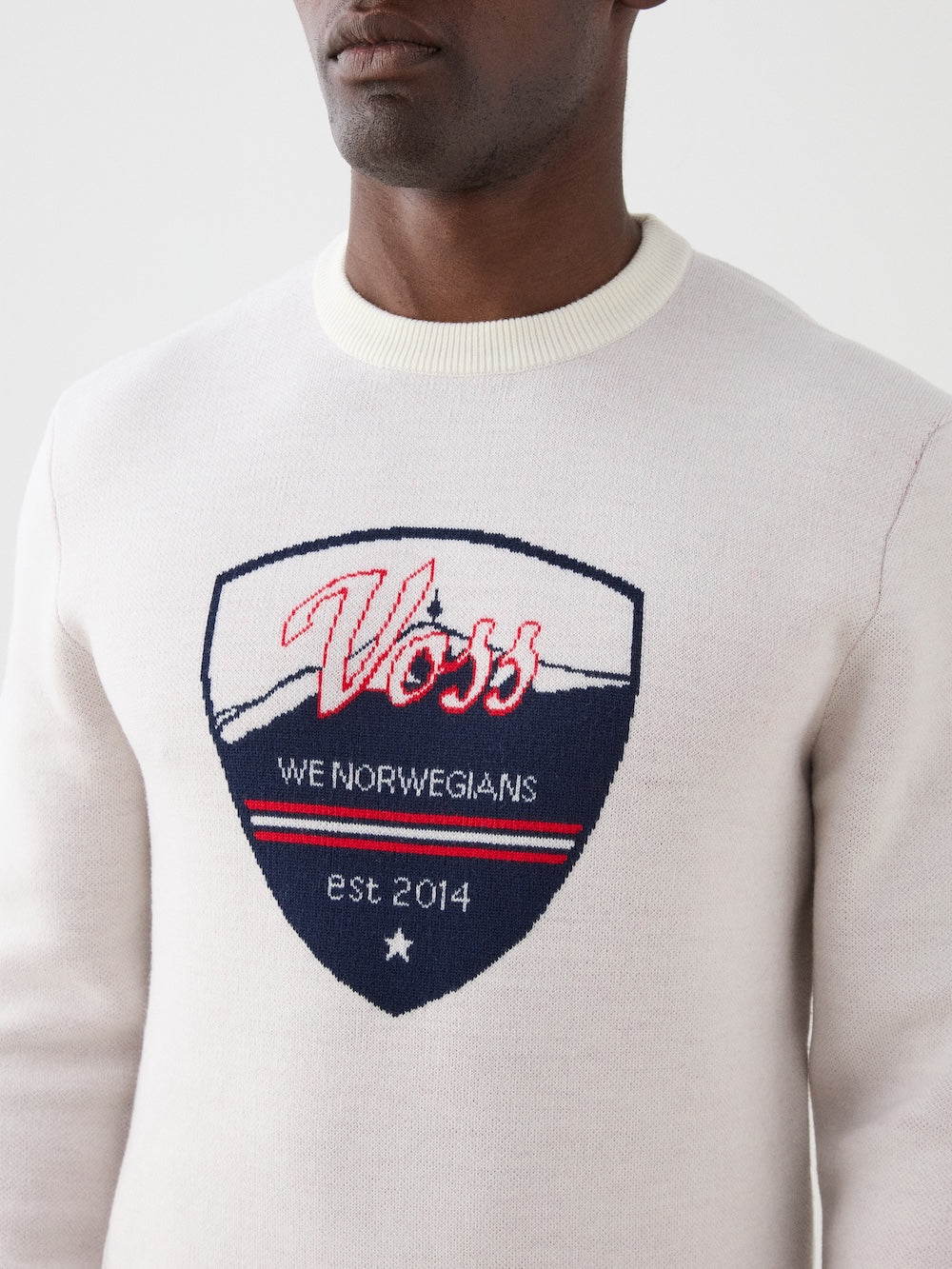 Someone wearing the We Norwegians AfterSki CrewNeck for men, a light-colored merino wool top featuring a shield patch with "Voss We Norwegians est. 2014" in red, white, and blue, plus a mountain outline and star.