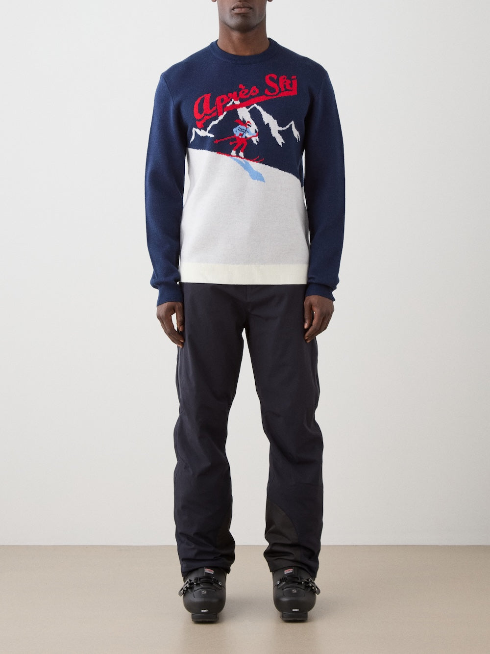 Someone stands indoors wearing black ski pants, black ski boots, and the We Norwegians AfterSki CrewNeck men sweater in navy blue with a white and red mountain graphic. The background is plain and minimal.