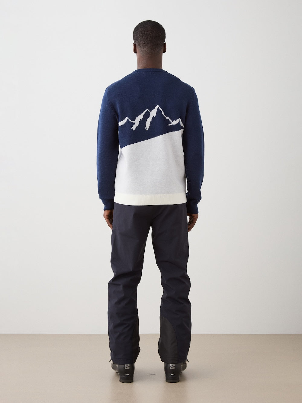 A person faces away, wearing dark pants and the We Norwegians AfterSki CrewNeck men’s sweater in navy with a white mountain design on the back, set against a plain, neutral background.