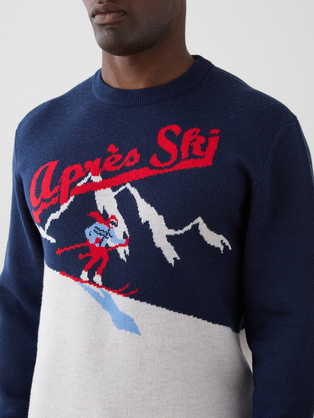 A man wears the We Norwegians AfterSki CrewNeck, a navy sweater with red “Après Ski” text and a skier graphic against snow-capped mountains.