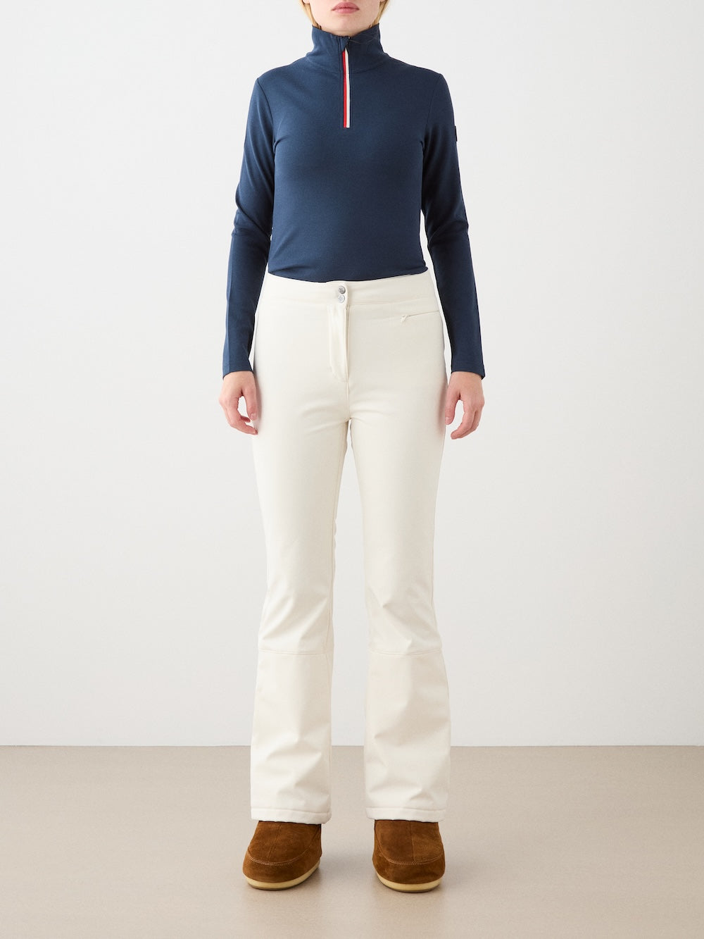 A person stands against a plain backdrop wearing the We Norwegians Bibi ZipUp Solid women’s top in navy, paired with off-white pants and brown boots—the perfect ski trip base layer. Their arms rest at their sides, head cropped out of frame.
