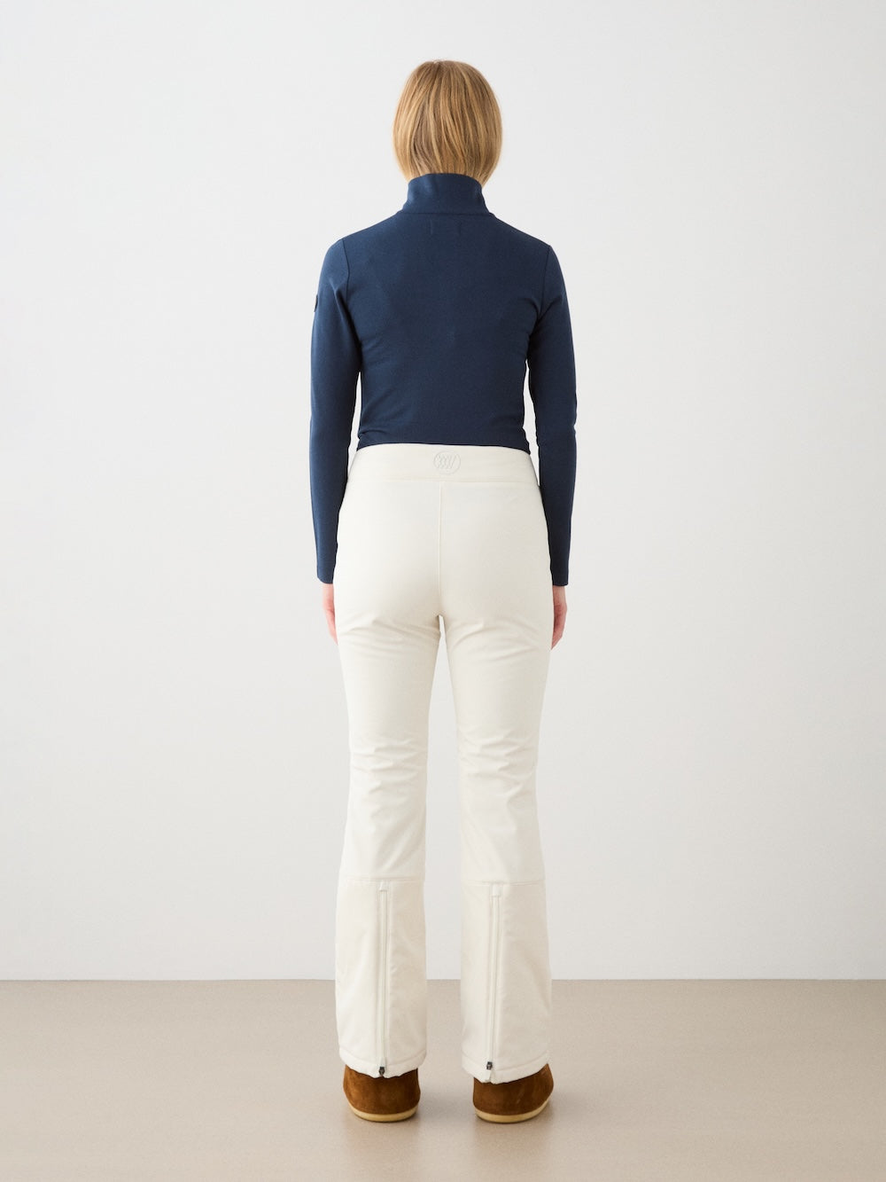 A person faces away from the camera, wearing the We Norwegians Bibi ZipUp Solid women navy Merino wool top, white pants, and brown boots against a plain white wall and beige floor.