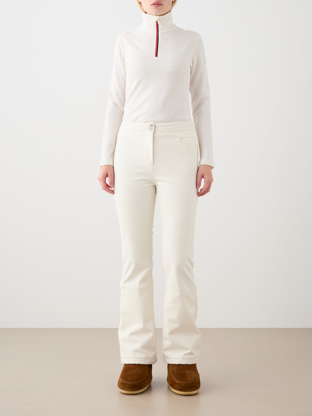 A person stands against a plain background wearing the We Norwegians Bibi ZipUp Solid women’s off-white Merino wool turtleneck with a red and black collar stripe, matching off-white pants, and brown boots—perfect for ski layering. Their face is not visible.