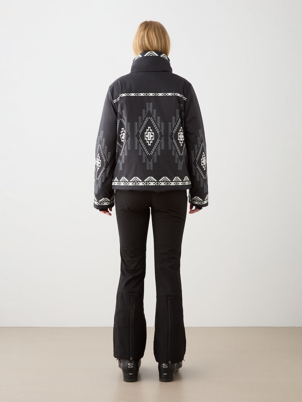 A woman with light brown hair faces away, wearing the We Norwegians Pioneer Ski Jacket for women—black with white geometric patterns—paired with black flared pants and shoes, set against a plain background.