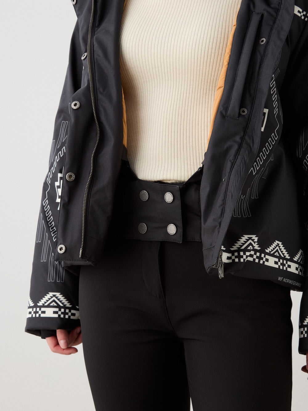 A woman wears the We Norwegians Pioneer Ski Jacket unzipped over a cream ribbed sweater and high-waisted black pants with a wide, double-buttoned waistband, showcasing her stylish layered look.