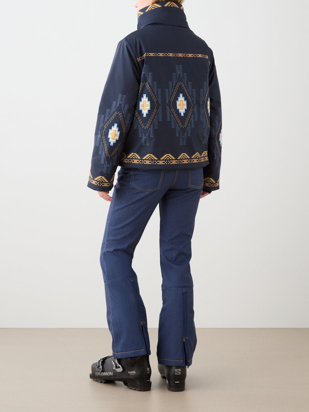 Indoors, a person faces away wearing the We Norwegians Pioneer Ski Jacket for women, featuring navy blue color and geometric designs inspired by Native American patterns, along with blue flared pants and black ski boots. Neutral tones fill the space.