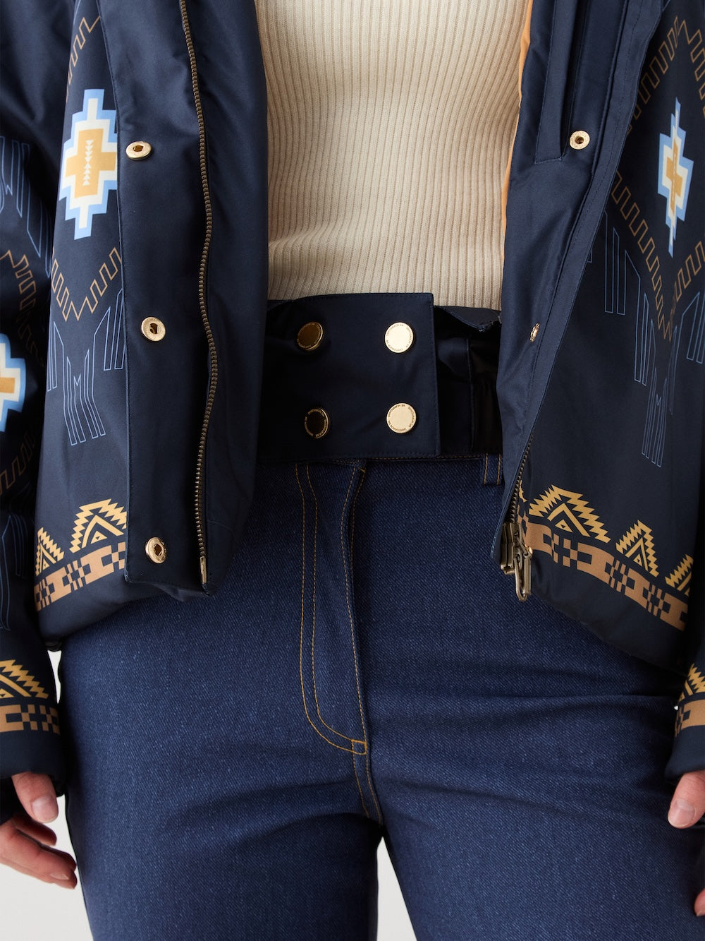 A person wearing a cream ribbed sweater, high-waisted blue jeans with yellow stitching, and a We Norwegians Pioneer Ski Jacket for women featuring geometric designs in blue, yellow, and beige.