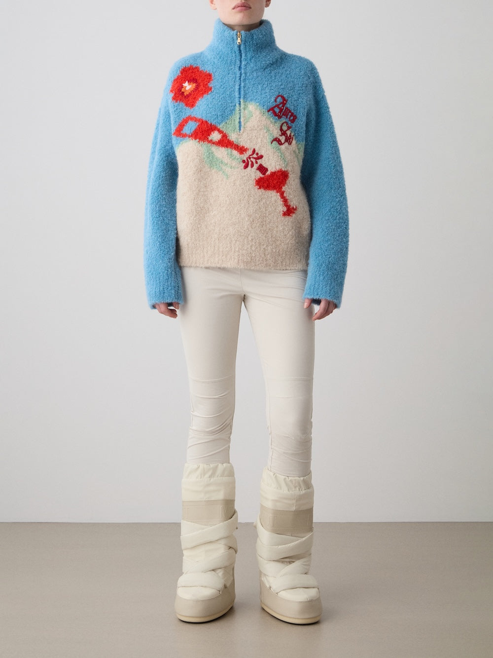 A person stands indoors in cream pants and chunky winter boots, wearing the limited edition WN x Markarian Bouclé Sweater for women by We Norwegians—a fuzzy blue and beige Italian bouclé knit featuring a colorful abstract design. A plain background is visible.