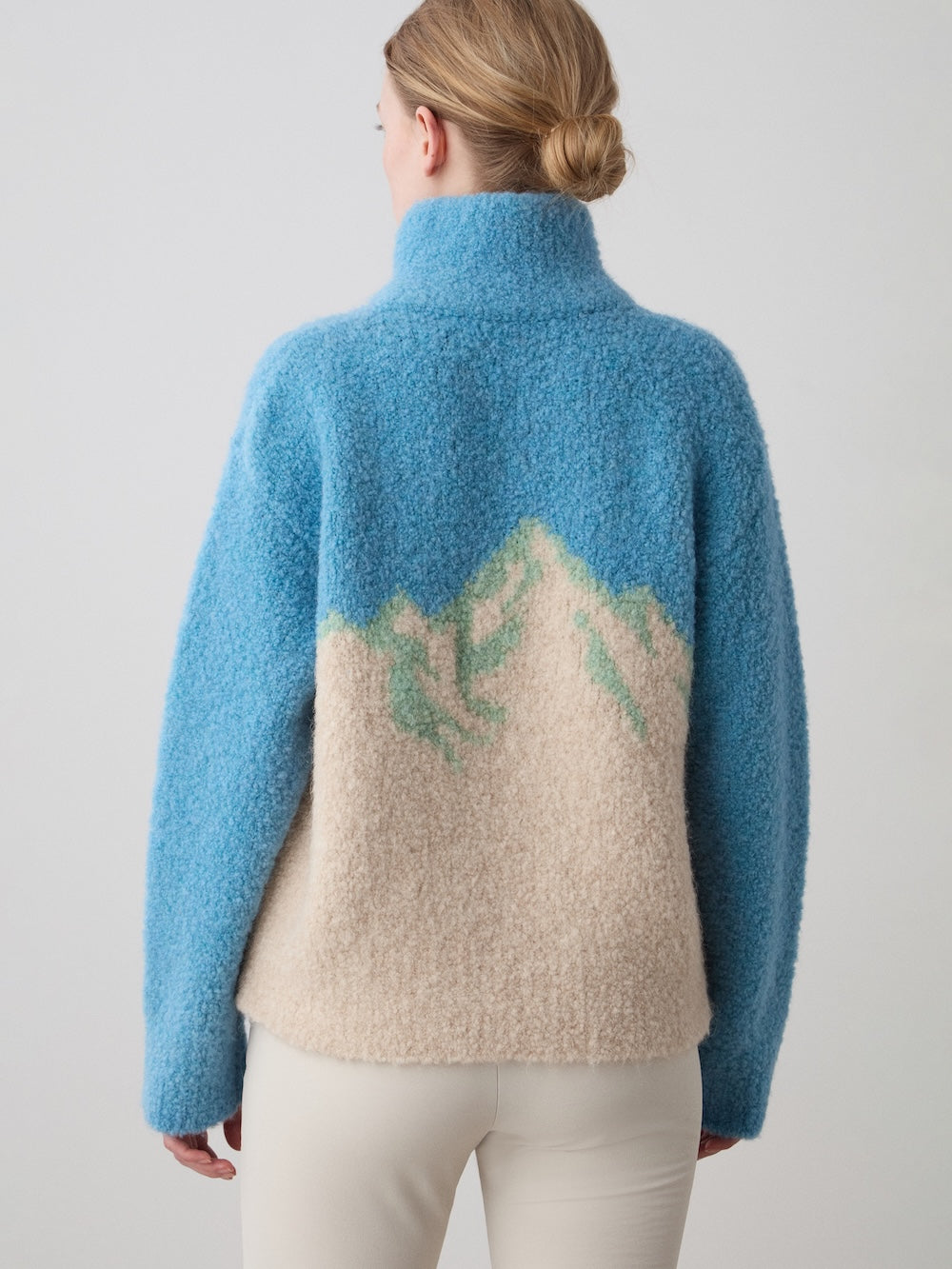 A woman with blonde hair in a low bun wears the WN x Markarian Bouclé Sweater for women by We Norwegians, featuring a blue and beige mountain-inspired design, paired with light pants. She faces away against a plain background.