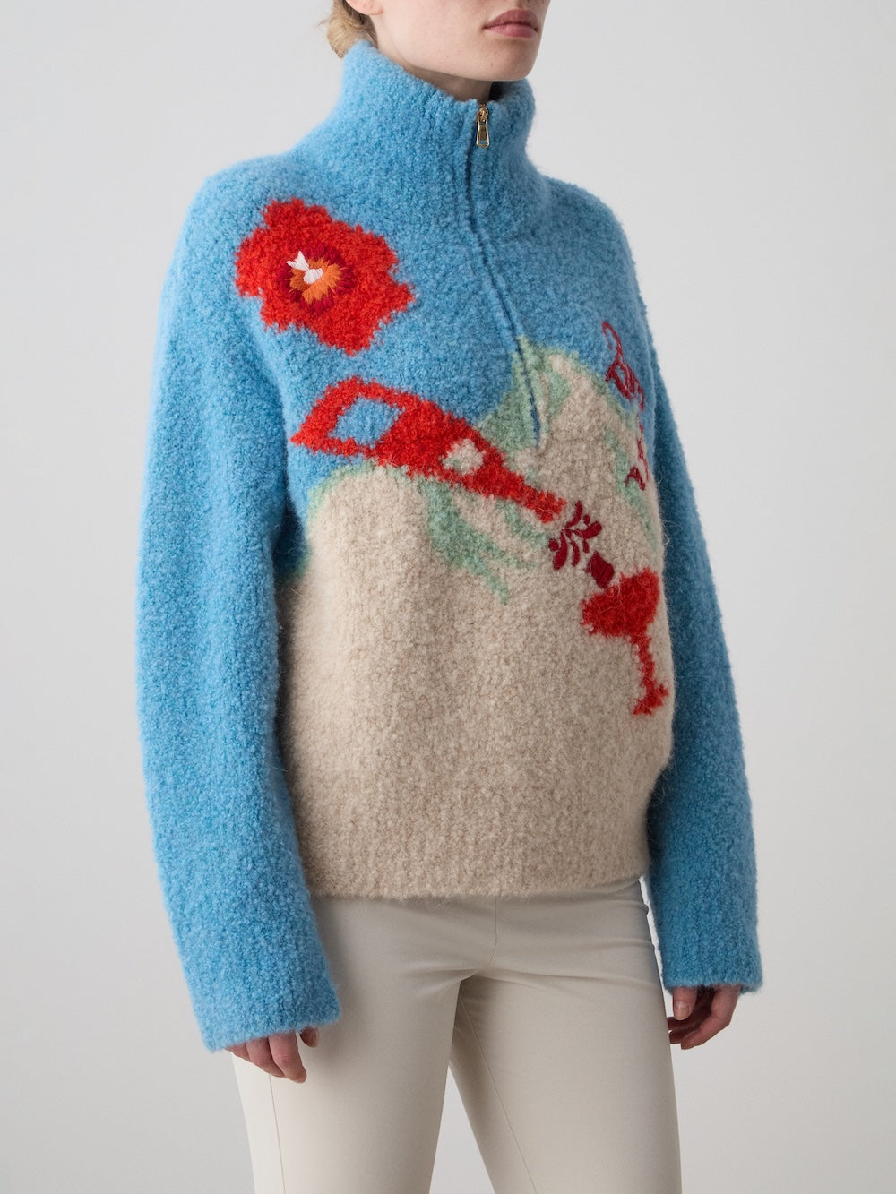 A woman wears the We Norwegians WN x Markarian Bouclé Sweater, featuring fuzzy blue and beige Italian bouclé yarn, a high collar, zipper, and colorful abstract floral designs on the front, paired with off-white pants.