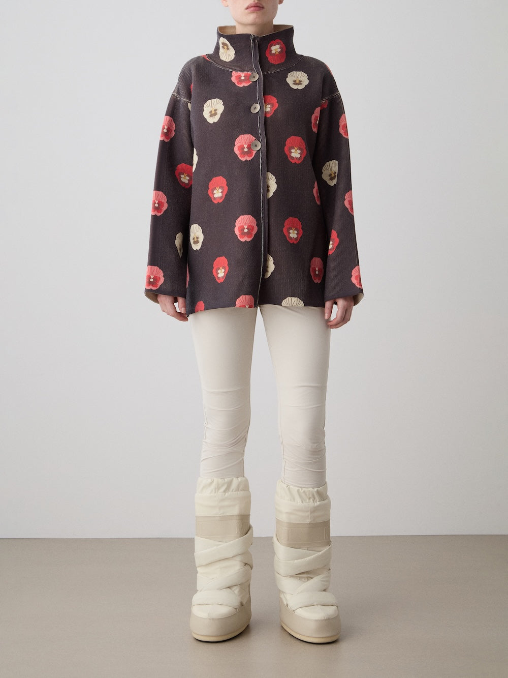 A person stands against a plain background wearing the oversized, limited edition We Norwegians WN x Markarian Jacket for women with a red and beige floral pattern, cream fitted pants, and chunky cream winter boots. Their face is not shown.