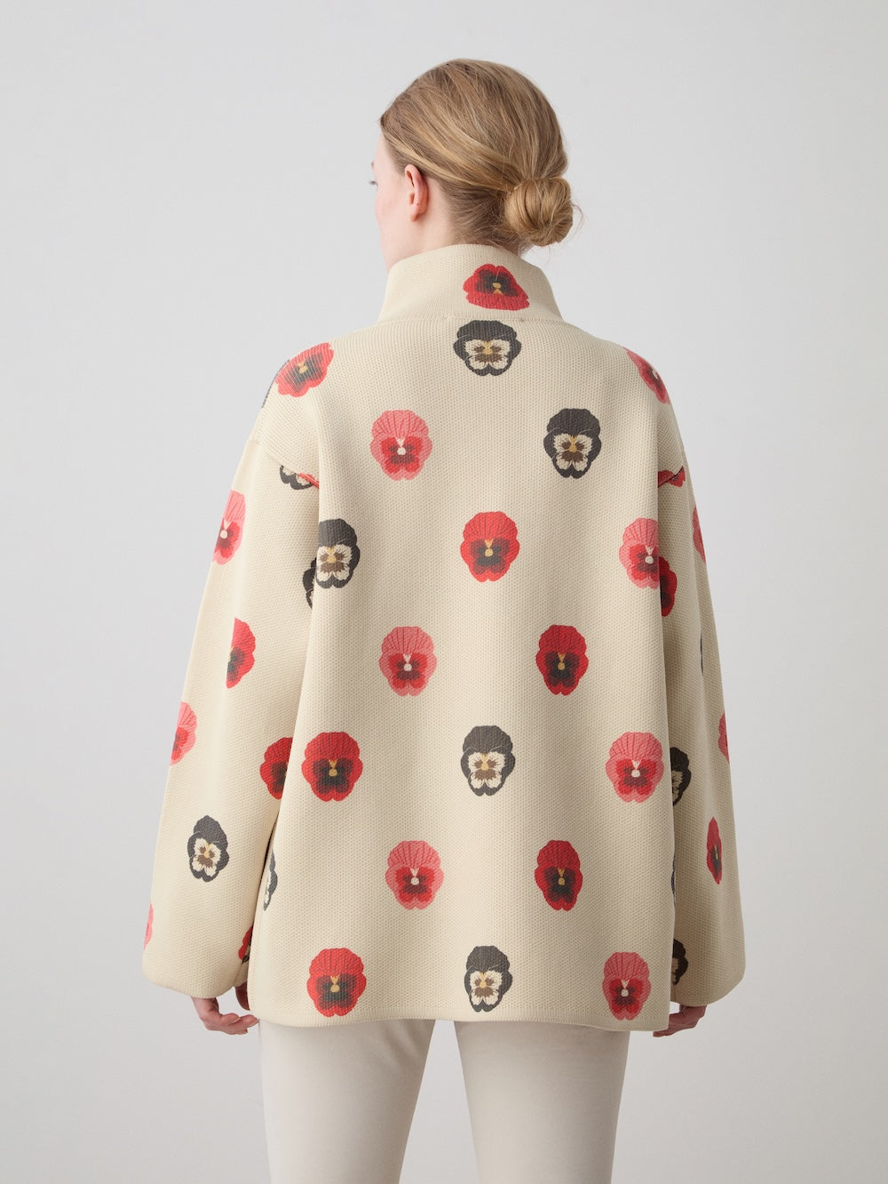 A woman with blonde hair in a low bun wears the WN x Markarian Jacket by We Norwegians, featuring digitally printed wool with red and dark brown flower faces, standing against a plain light background and facing away from the camera.