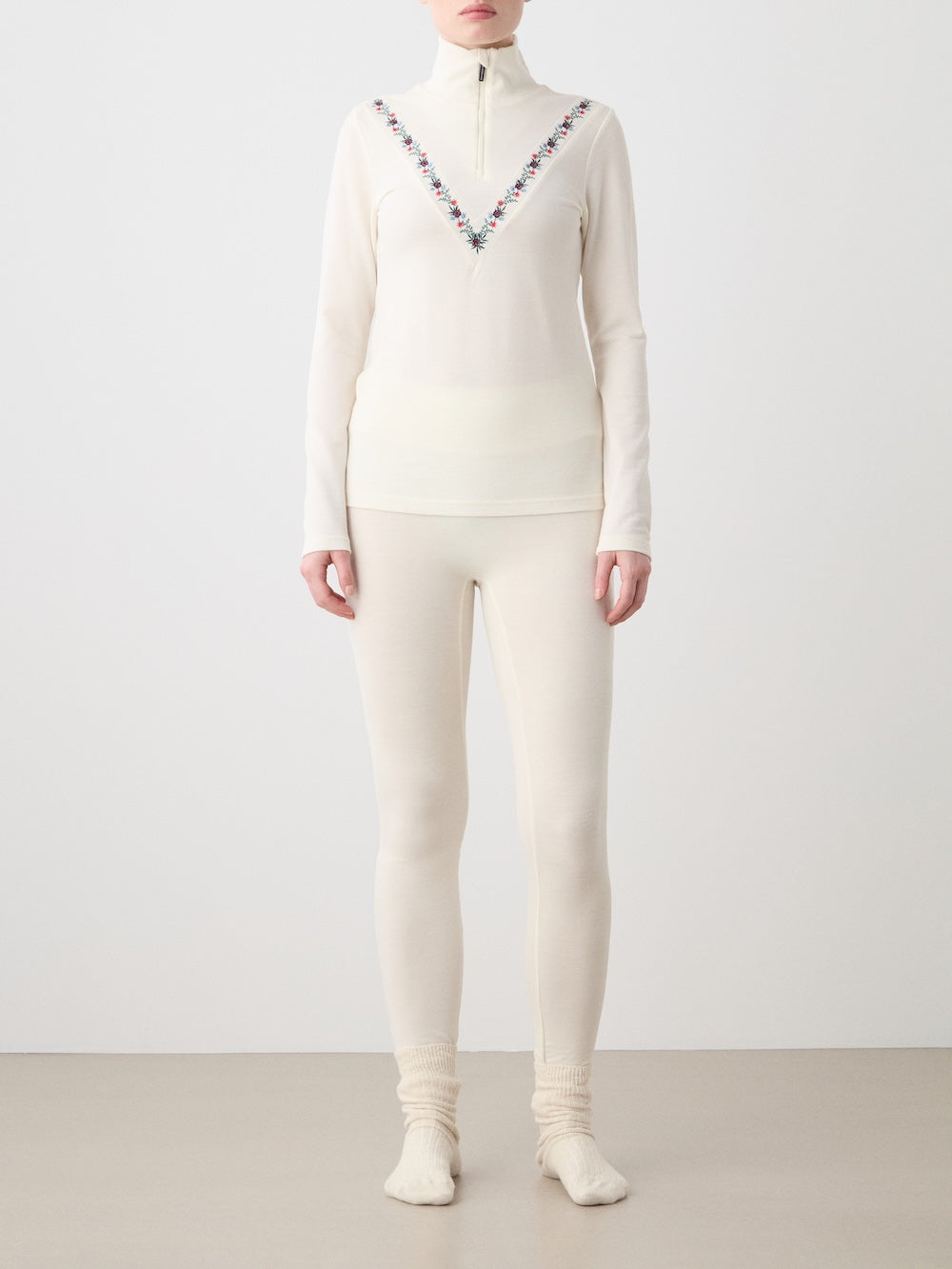 Against a plain background, a person wears the We Norwegians WN x Markarian ZipUp women—an off-white, merino wool, long-sleeve base layer with a half-zip and V-shaped embroidery—plus matching leggings and cream socks.
