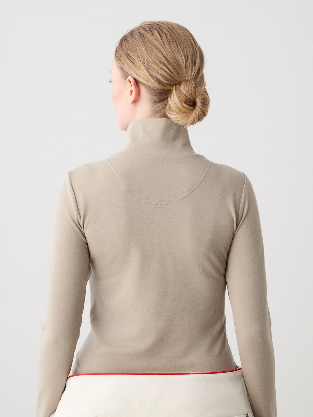 A person with blond hair in a low bun is seen from behind, wearing a beige turtleneck and the We Norwegians WN x Markarian ZipUp women base layer. The background is plain and light-colored.