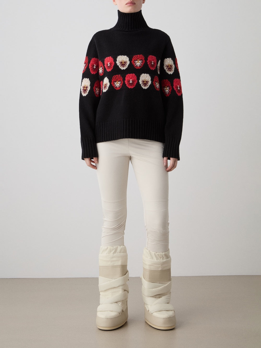 Against a plain background, a person models the We Norwegians WN x Markarian Sweater women featuring red and white flower patterns, paired with cream pants and chunky winter boots. Their face is not visible.