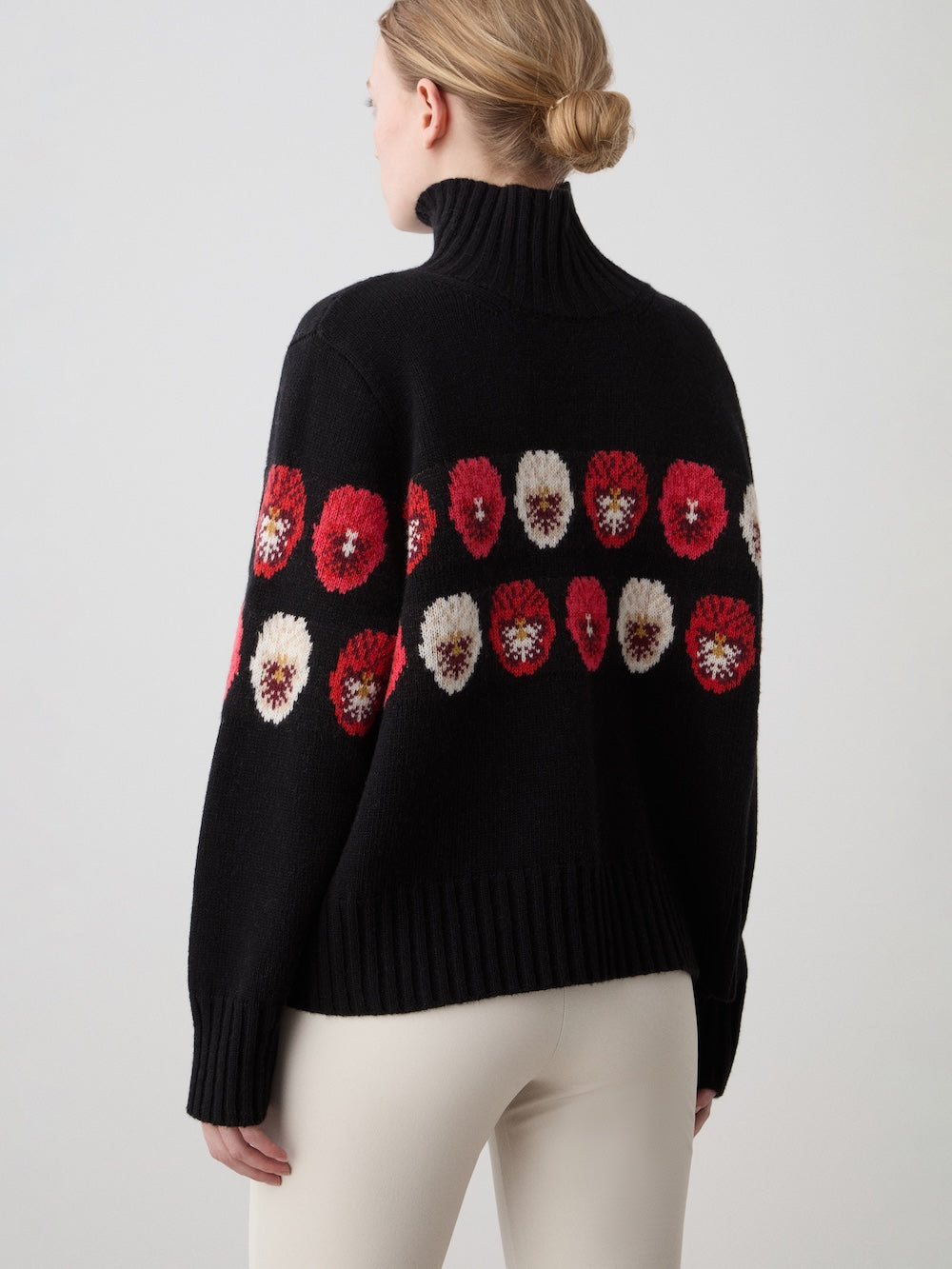 A blonde-haired woman with a low bun wears the limited edition WN x Markarian Sweater by We Norwegians, featuring black Fair Isle with red and white flowers, paired with light beige pants, against a plain background.