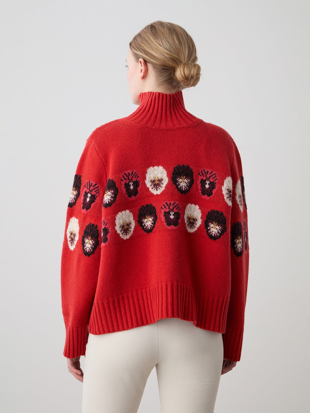 A person with blonde hair in a low bun, facing away, wears cream pants and the limited edition WN x Markarian Sweater women by We Norwegians—red with a multicolored abstract face pattern across the back and sleeves.