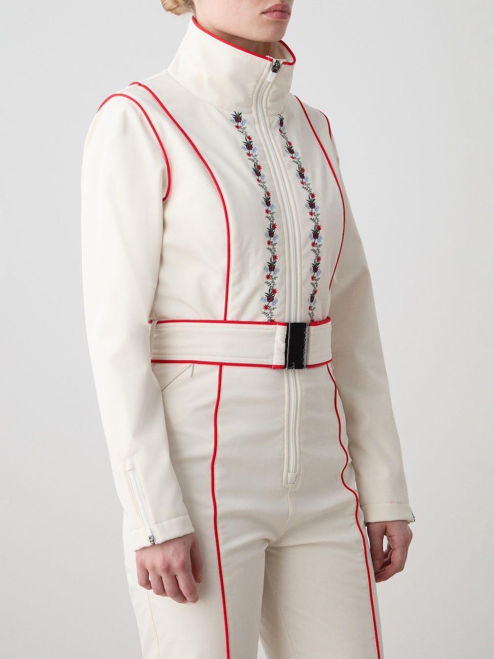 A person wearing the We Norwegians WN x Markarian Ski Jumpsuit for women—a fitted white ski suit with red piping, floral embroidery along the front zipper, matching belted waist, long sleeves, and a high collar. Face is partially out of frame.