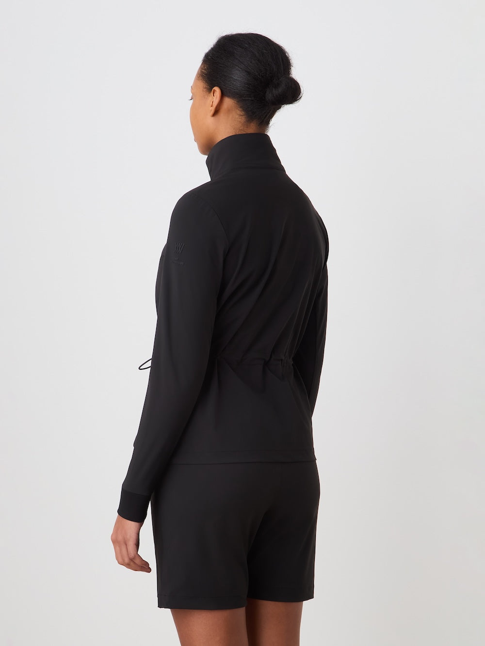 A woman with dark hair in a bun is seen from behind wearing the We Norwegians Albatross Jacket Women—black, stretch zip-up—and black shorts, set against a plain light background.