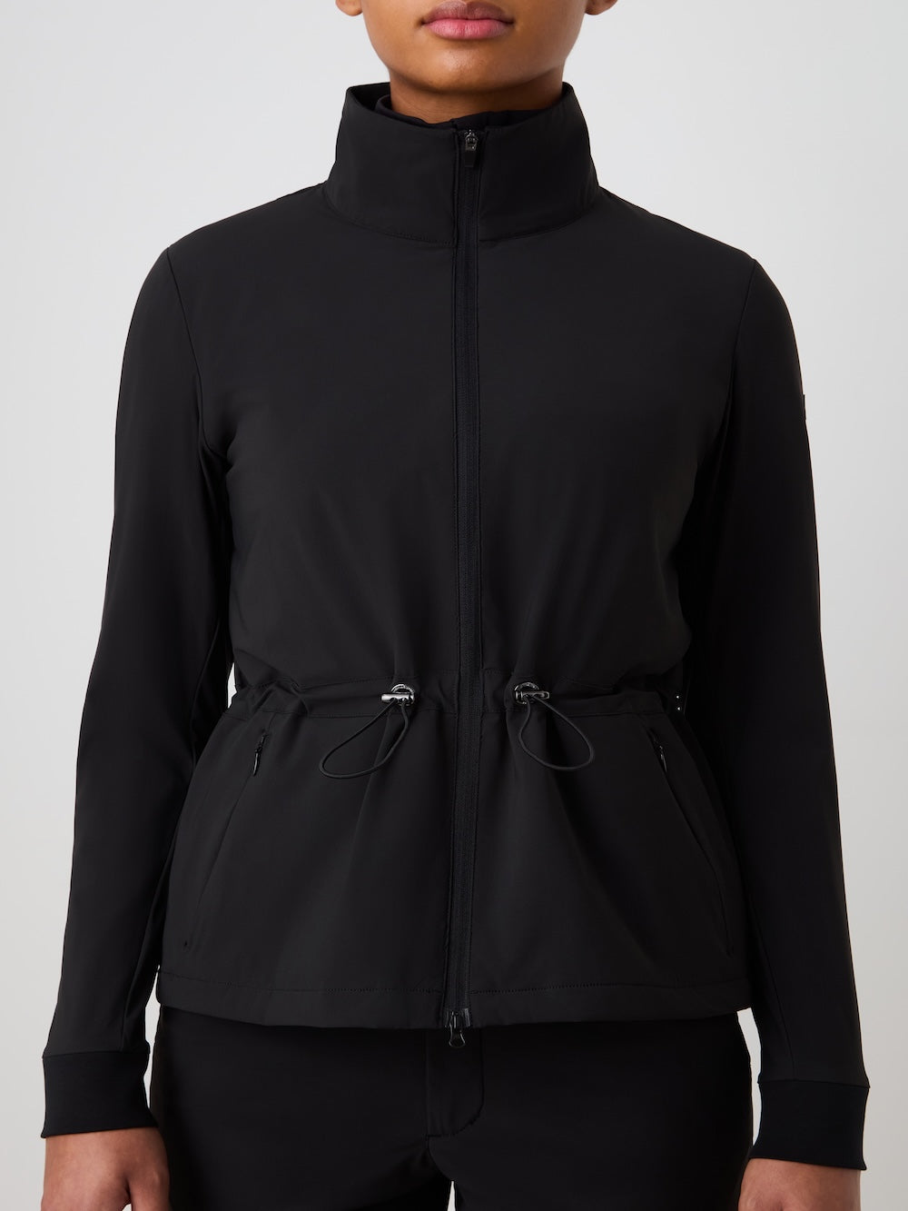A woman wears the We Norwegians Albatross Jacket, a black, lightweight golf jacket featuring a high collar, stretch fabric, and adjustable waist drawstrings, standing against a plain light background.
