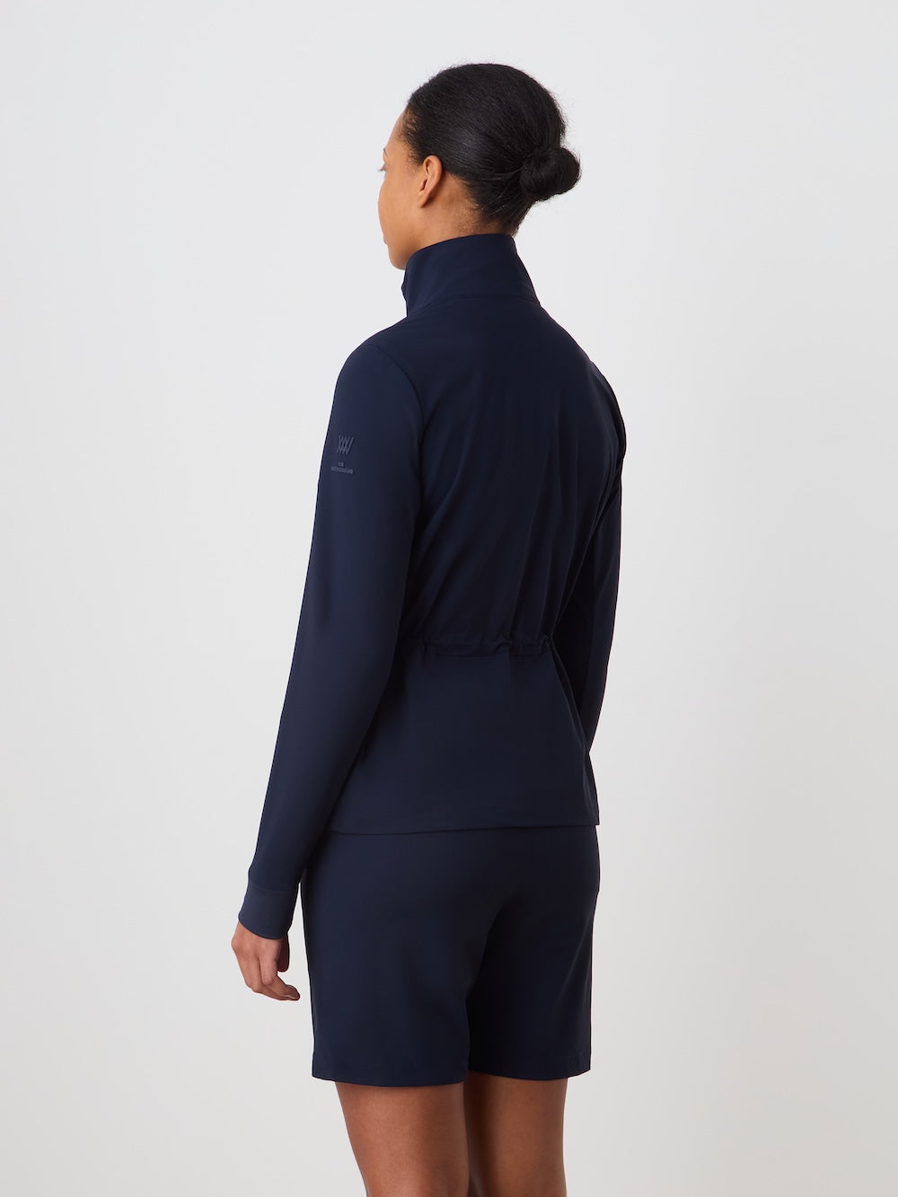 Seen from behind, a woman with dark hair in a low bun wears the We Norwegians Albatross Jacket Women—a navy lightweight golf jacket with high collar—and matching navy shorts, standing against a plain light background.